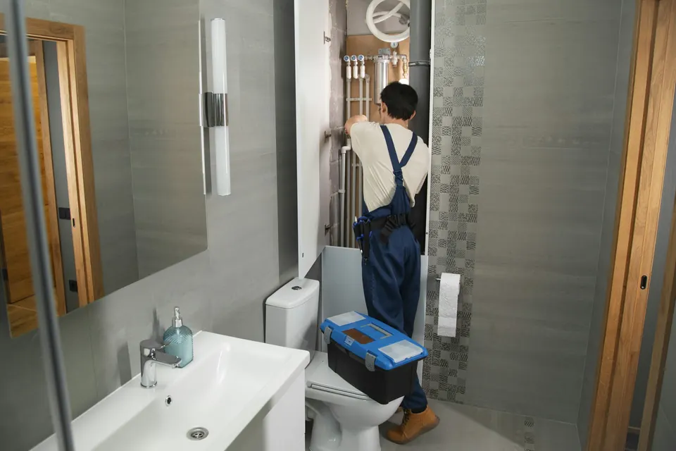 A plumber in work overalls with a toolbox, working on pipes in a bathroom
